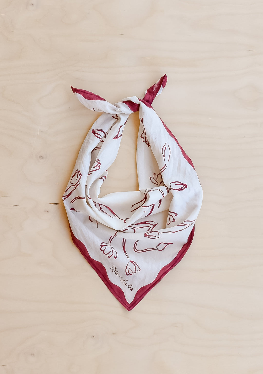 Close-up of a TBCo x Harlie Briggs Artist Series cotton triangle neck scarf in white with red floral line art, featuring the TBCo x Harlie logo, against a soft wooden  background.

 