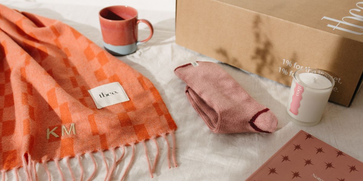 TBCo personalised embroidery orange check lambswool scarf with ‘KM’ initials, styled with pink merino wool socks, a ceramic TBCo mug, candle, notebook and branded gift box – a cosy, sustainable gifting set.