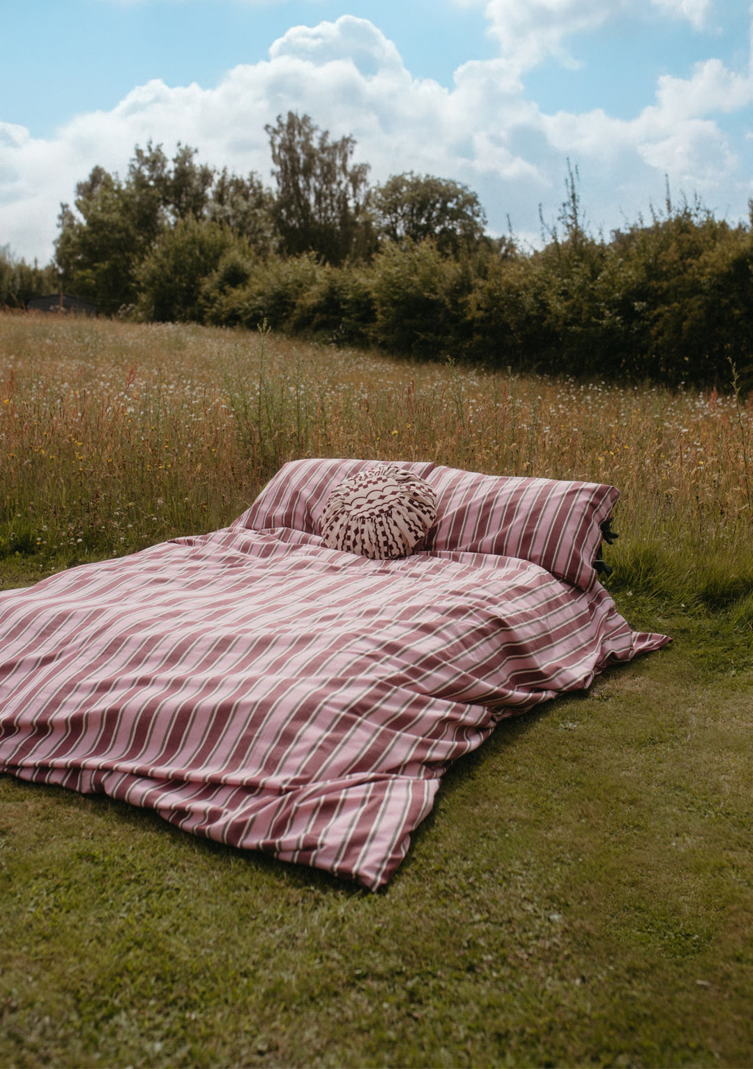 Cotton Duvet Cover in Pink Stripe by TBCo displayed outdoors on grass with pillows and nature background.