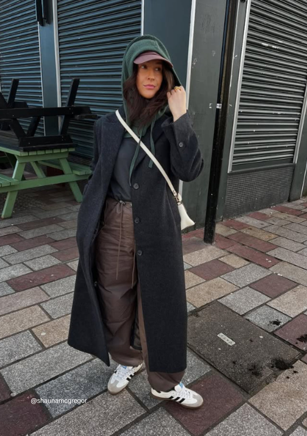 Model wearing TBCo Merino Hood in Green with black winter coat, brown trousers and white trainers on urban street