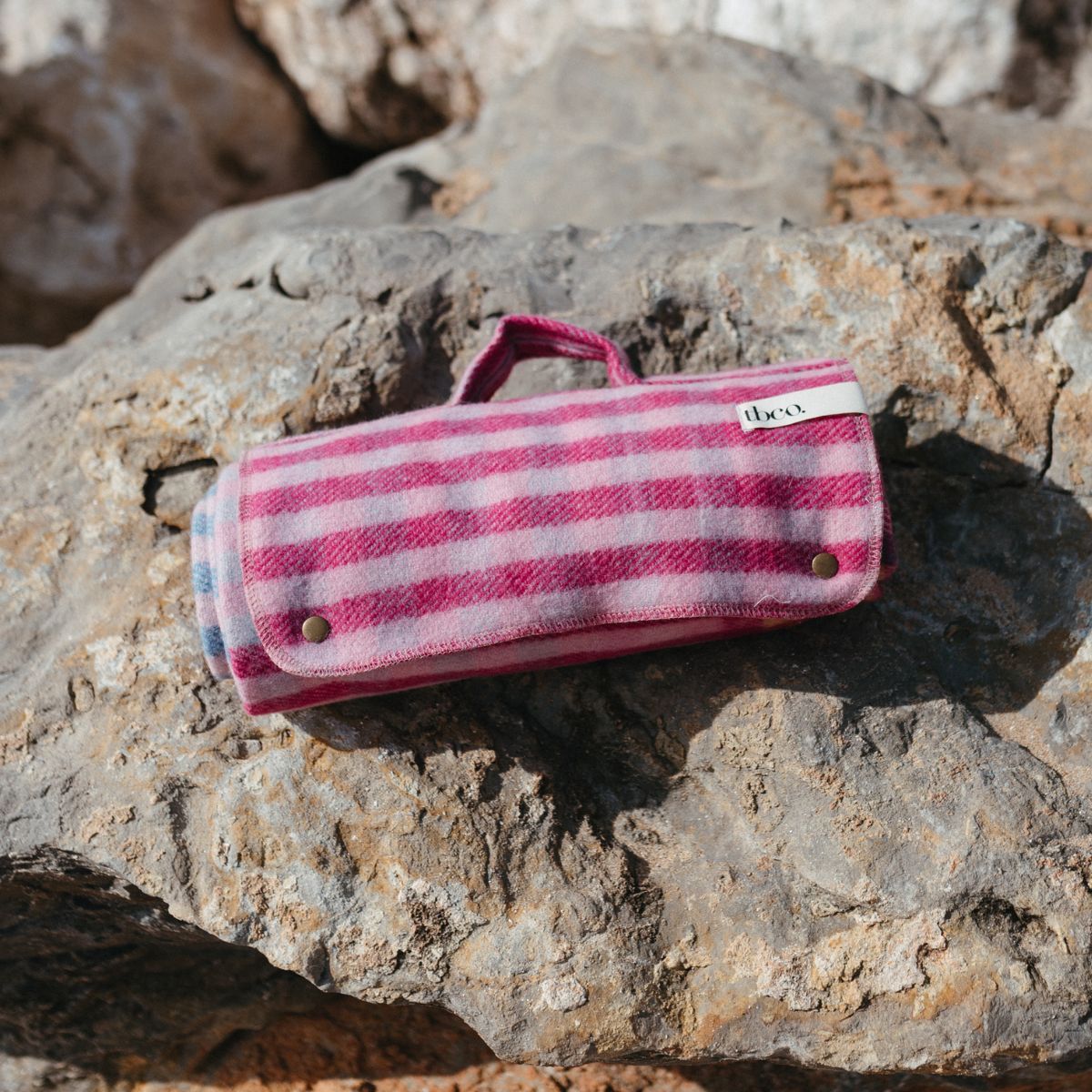 A TBCo small picnic blanket in a pink check sits rolled up on a rocky beach.