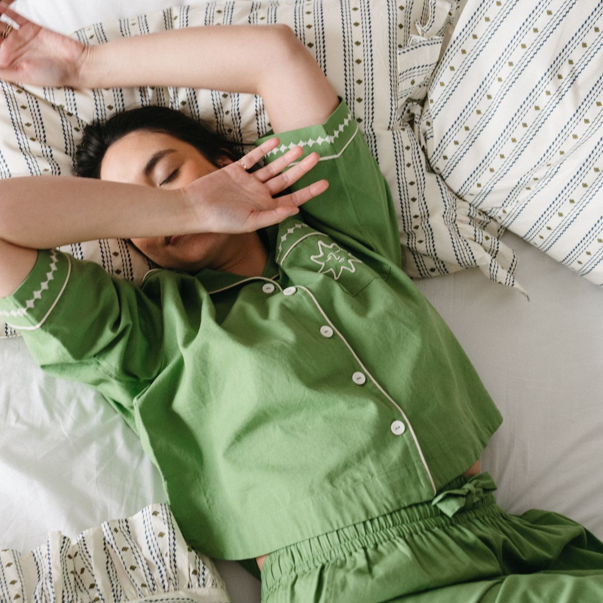 A model wearing green TBCo pyjamas lays across a bed made up with TBCo cream stripe bedding.