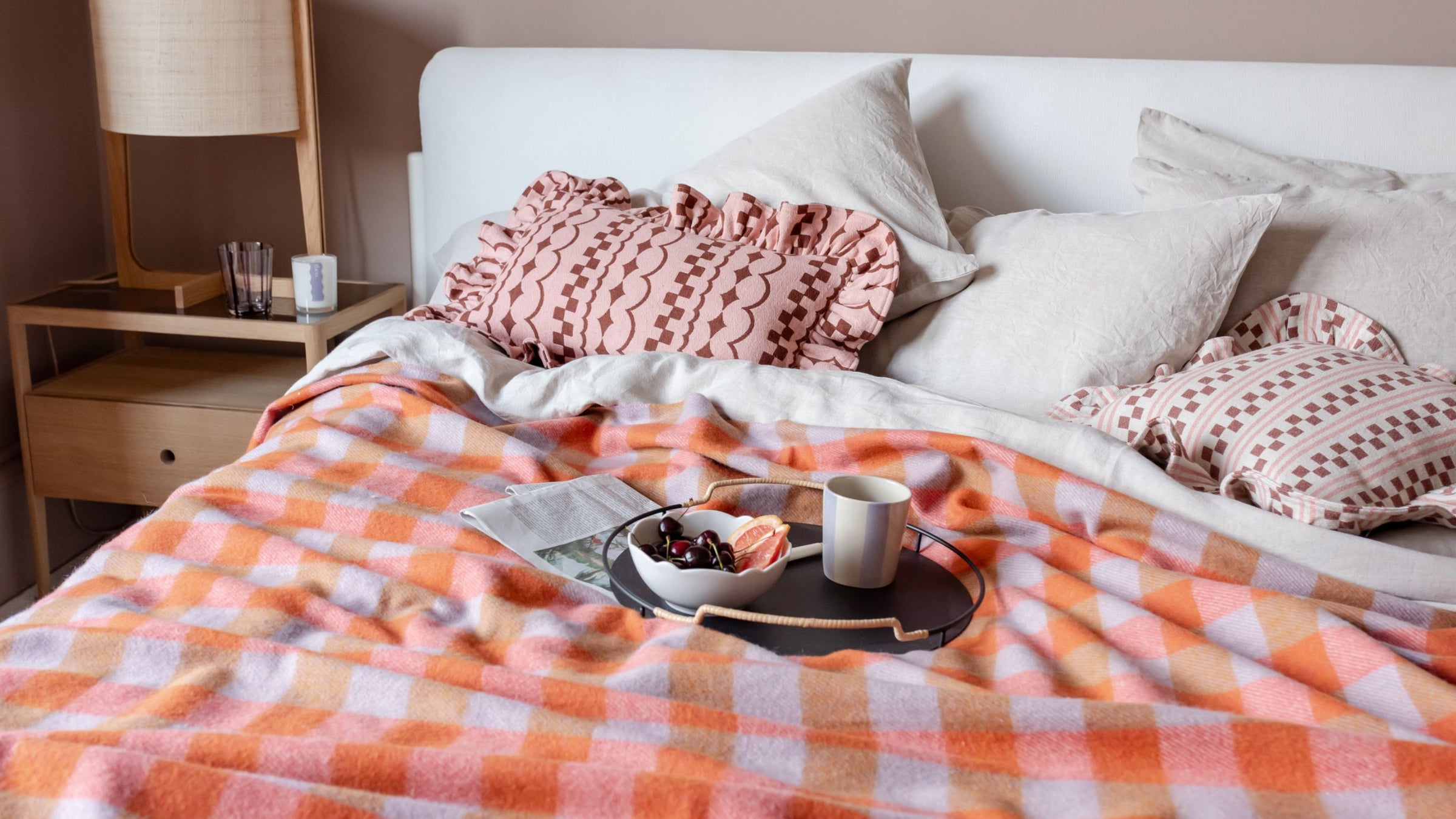 TBCo Recycled Wool Orange Ginga=ham blanket in a check pattern draped over a cosy bed with TBCo Cotton textured Pink  ruffle cushions. A perfect setting for relaxed mornings with coffee and pastries.
