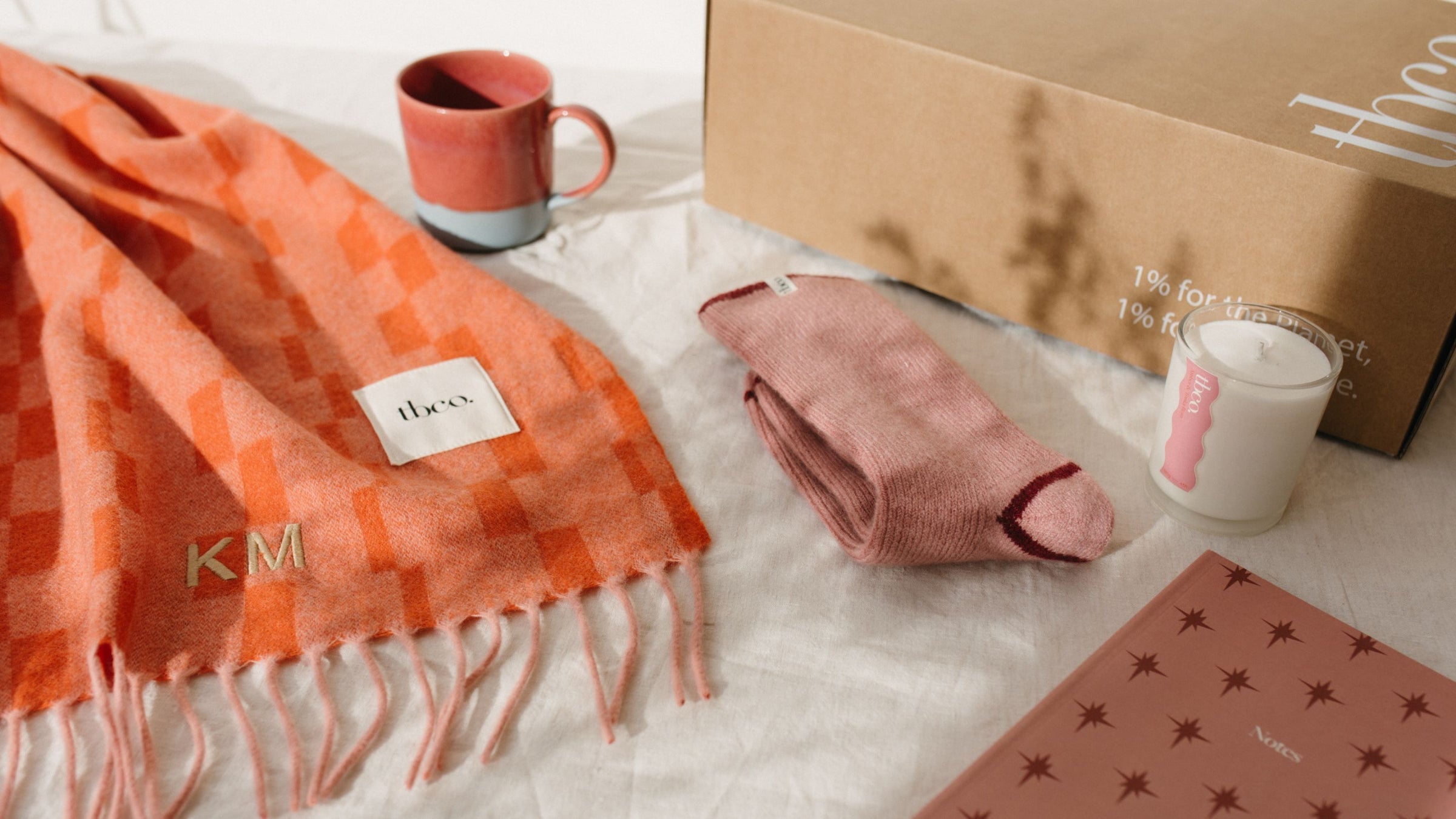 TBCo personalised embroidery orange check lambswool scarf with ‘KM’ initials, styled with pink merino wool socks, a ceramic TBCo mug, candle, notebook and branded gift box – a cosy, sustainable gifting set.