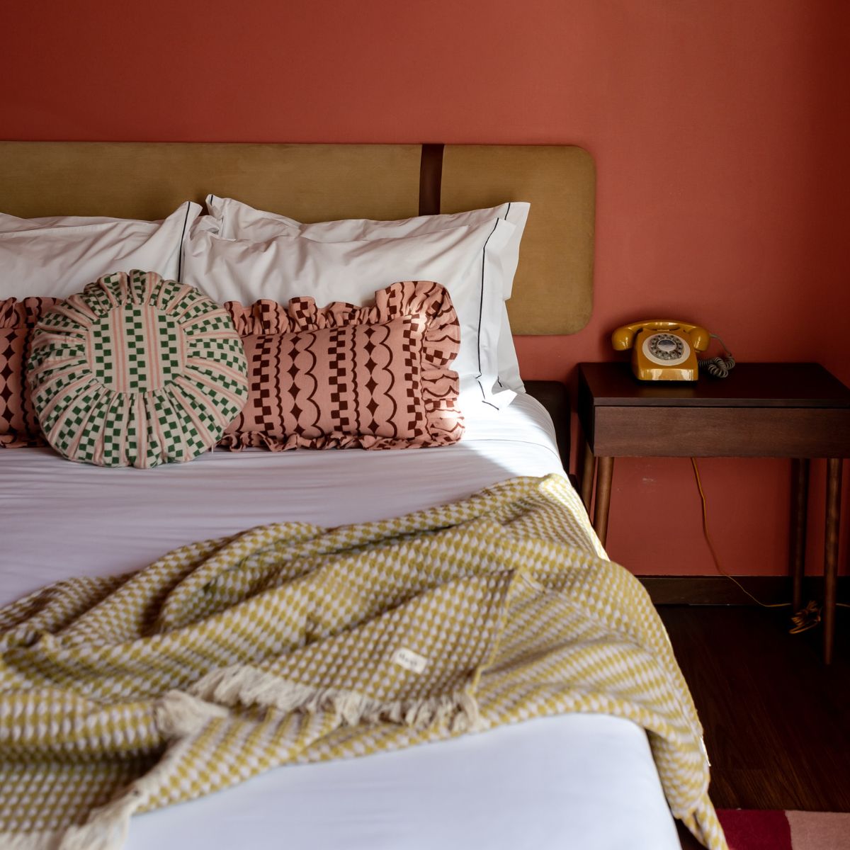 A bed with an olive green velvet headboard sits against a terracotta coloured wall. Plain white bedding is accessorized with two TBCo pink and burgundy frill cushions and a TBCo green cotton throw. 
