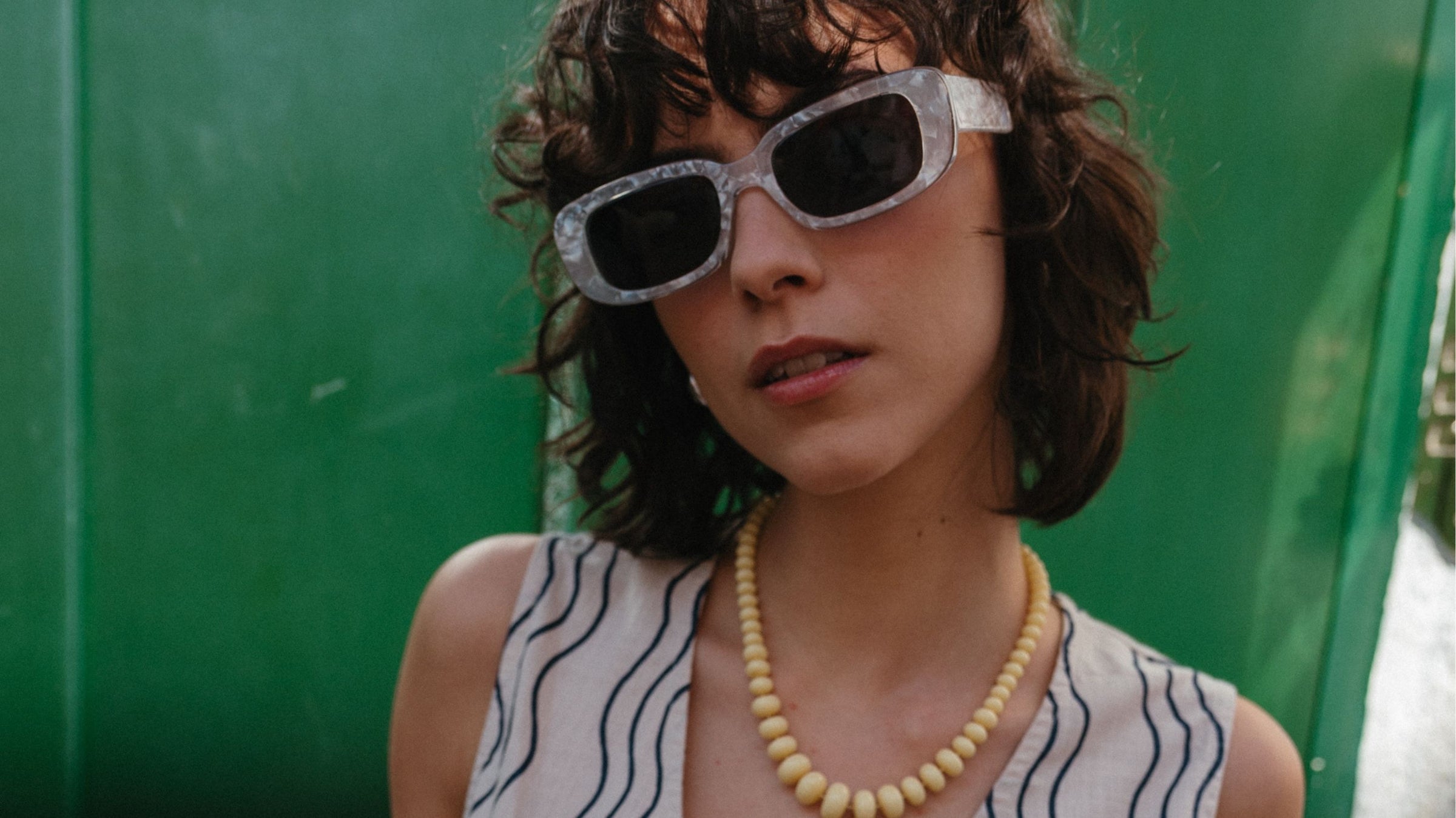 A model poses against a green door on a Lisbon street, wearing the TBCo blue wave trouser and vest set, accessorized with white sunglasses and yellow bead necklace.