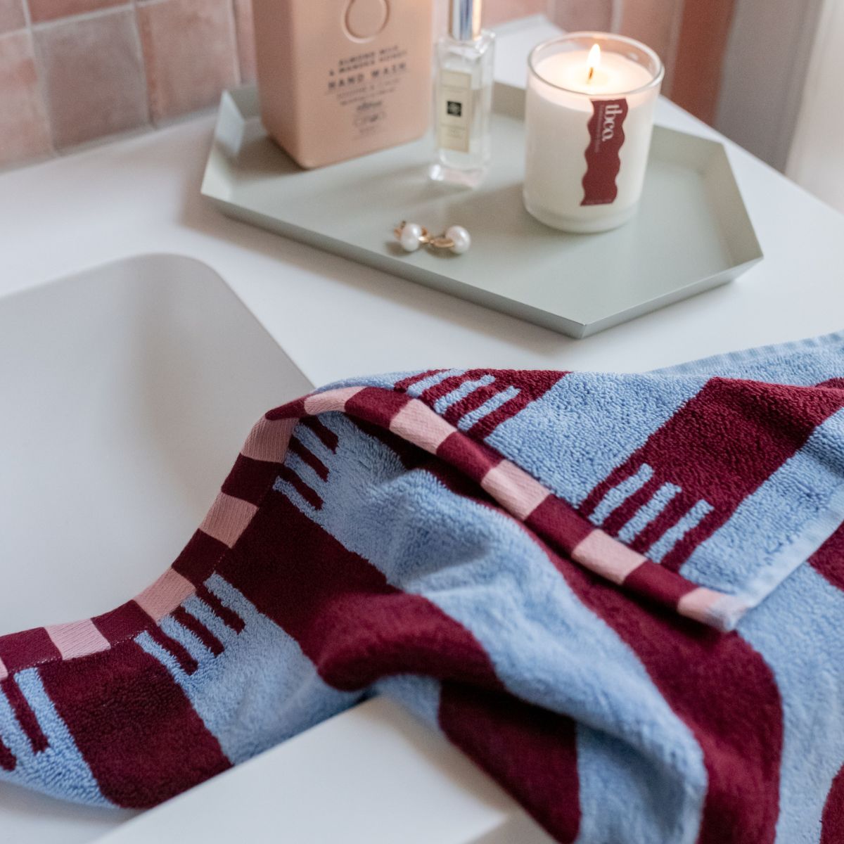 TBCo striped cotton towel in burgundy, sky blue, and soft pink elegantly draped over a bathroom sink. A luxurious and sustainable bath accessory styled alongside a TBCo lit candle, hand wash, and pearl earrings for a serene self-care moment