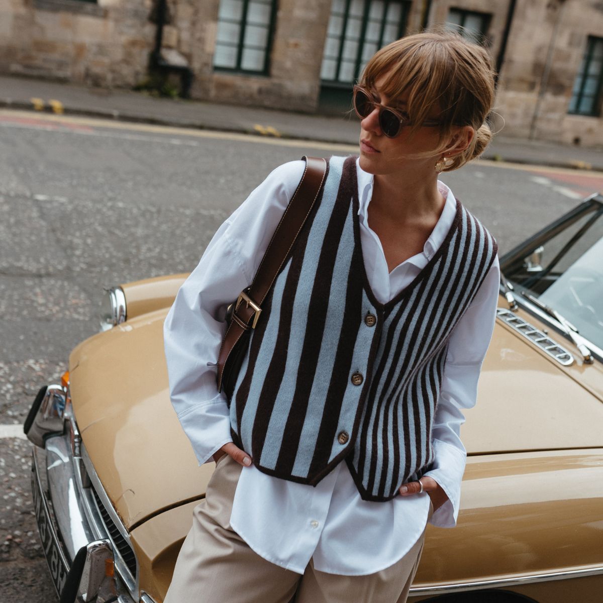Woman wearing a striped TBCo merino wool knit blue and brown vest over a crisp white shirt, paired with beige trousers and sunglasses, leaning against a vintage mustard-yellow car on a cobbled city street.