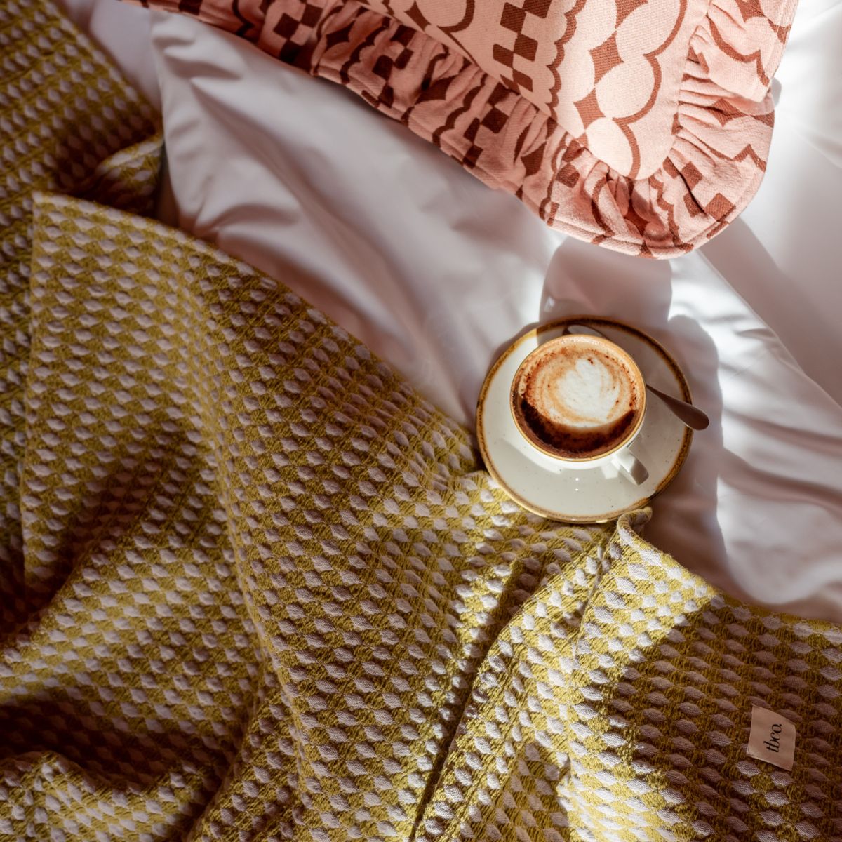TBCo Cotton Olive Argyle Throw on a bed with a Cotton Pink Ruffle Cushion with a geometric pattern, showing the perfect cosy moment with a cup of morning coffee.