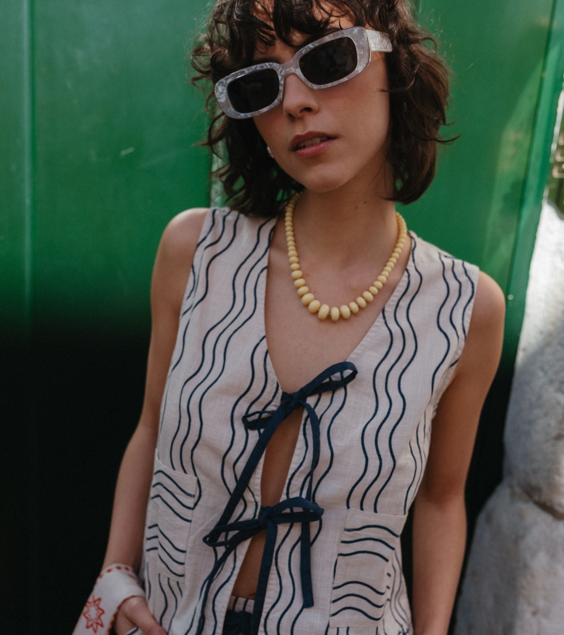 A model poses against a green door on a Lisbon street, wearing the TBCo blue wave trouser and vest set, accessorized with white sunglasses and yellow bead necklace.