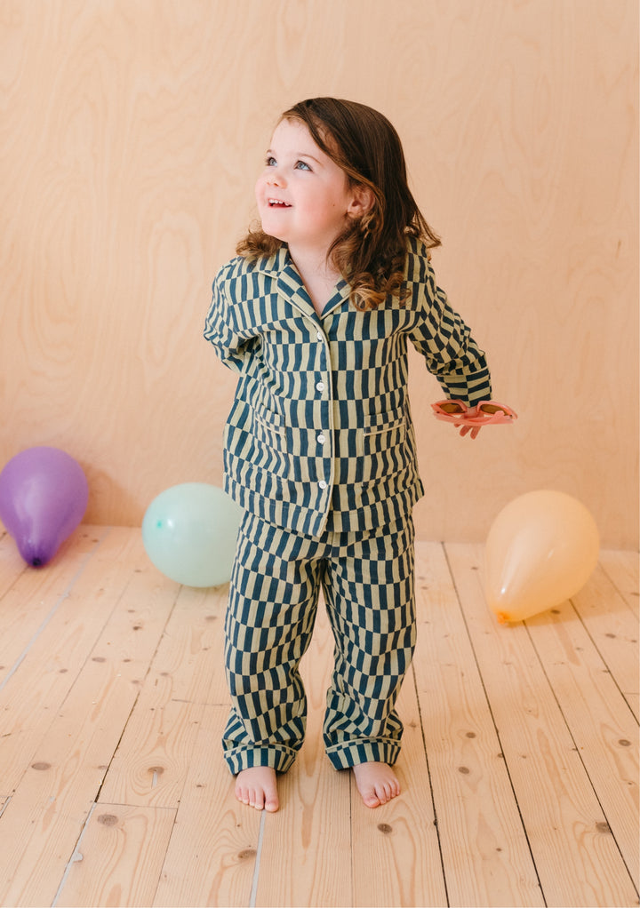 Young child wearing TBCo Cotton Kids Pyjamas in Green Checkerboard pattern, standing with colourful balloons on wooden floor