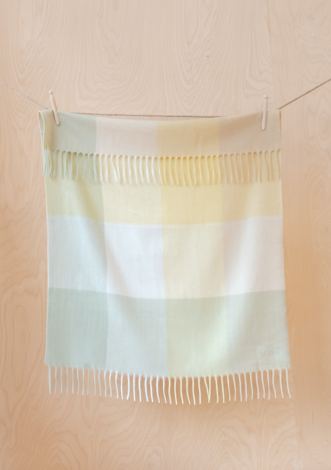 TBCo Lambswool Baby Blanket in Green Block Check pattern displayed with fringe detail, hanging on wooden background