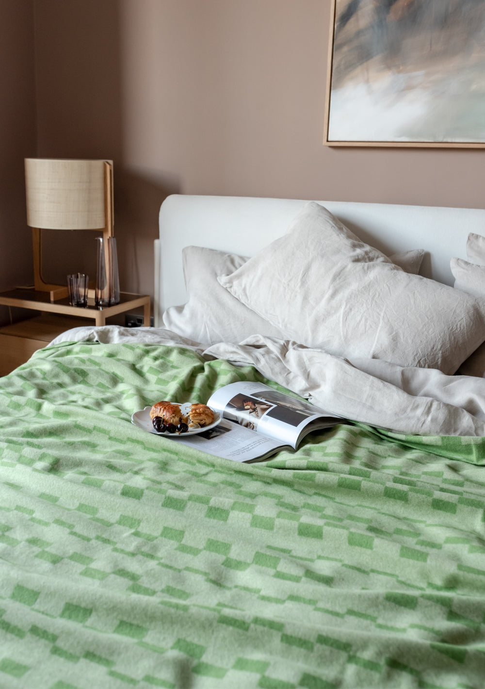 TBCo Lambswool Blanket in Green Checker Stripe draped over white bedding, styled with book and pastries on minimalist bed