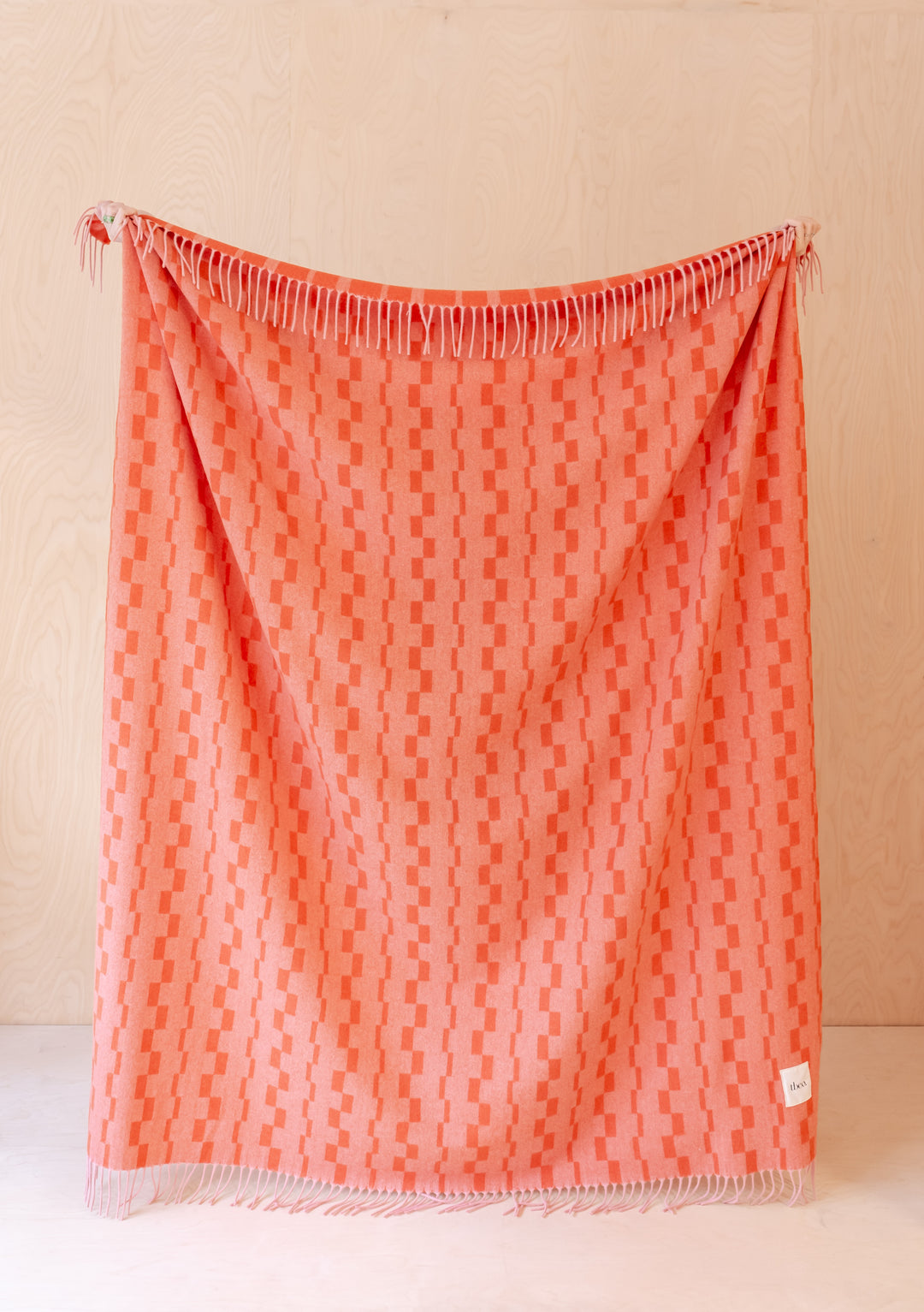 TBCo Lambswool Blanket in Tangerine Checker Stripe displayed with fringe detail against beige wall, showcasing geometric pattern