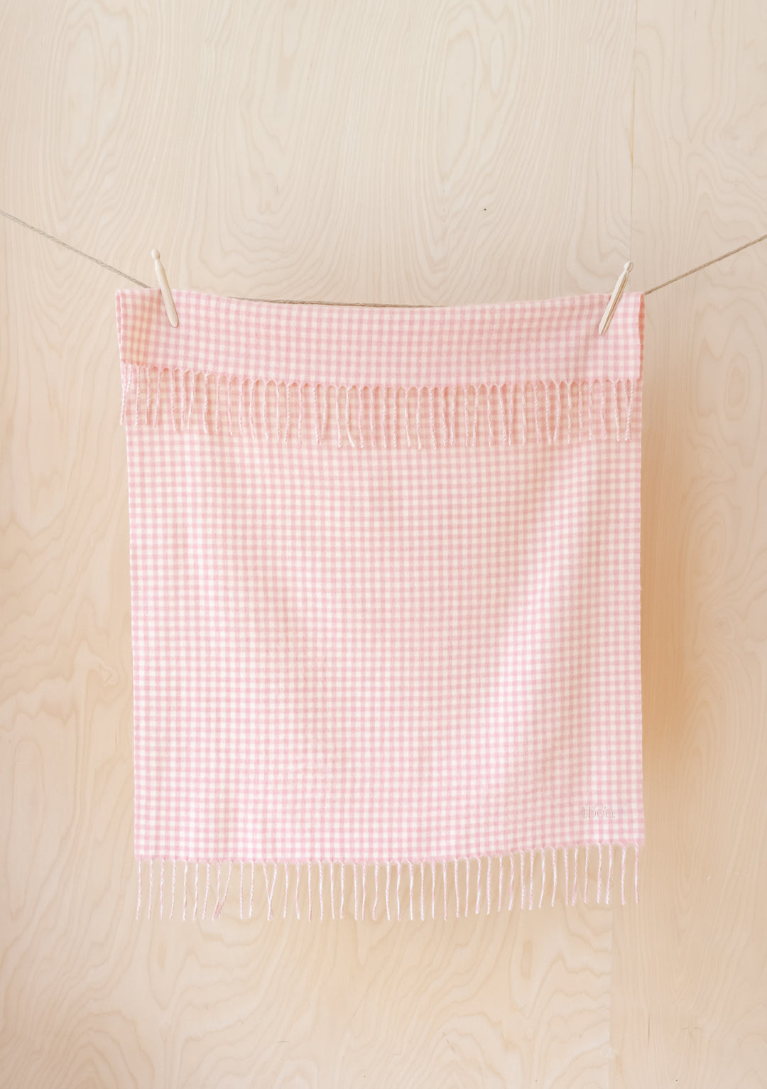 TBCo pink gingham lambswool baby blanket with fringe detail hanging on clothesline against cream wooden background