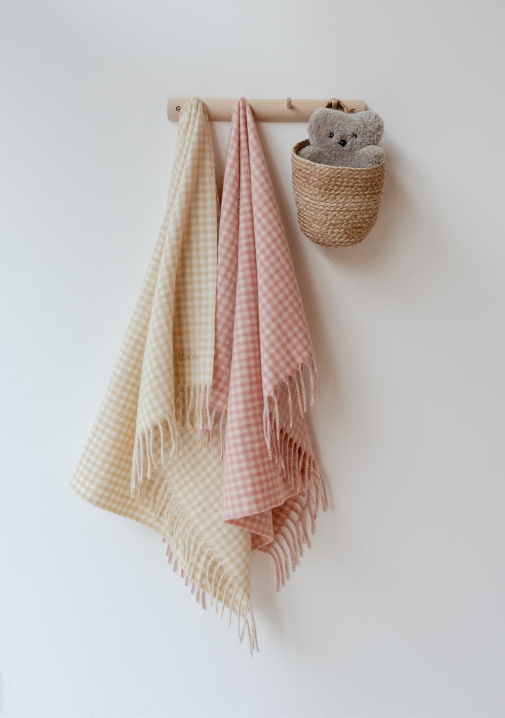 TBCo Lambswool Baby Blanket in pink and cream gingham pattern hanging on wooden peg rail with woven basket and soft toy