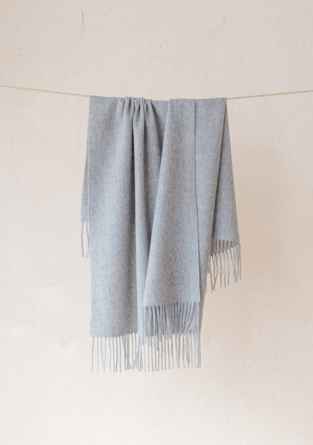 TBCo Lambswool Baby Blanket in Light Grey Melange hanging on rope with fringed edges against cream wall