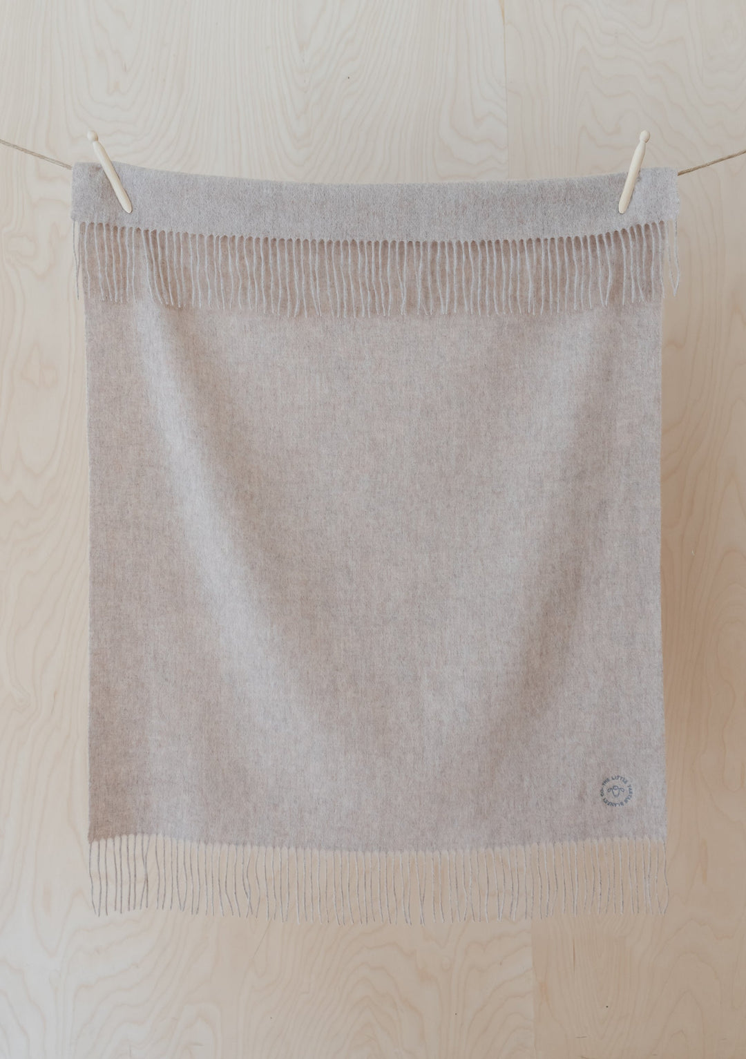 TBCo Lambswool Baby Blanket in Oatmeal Melange with fringed edges hanging on wooden pegs against light wooden background