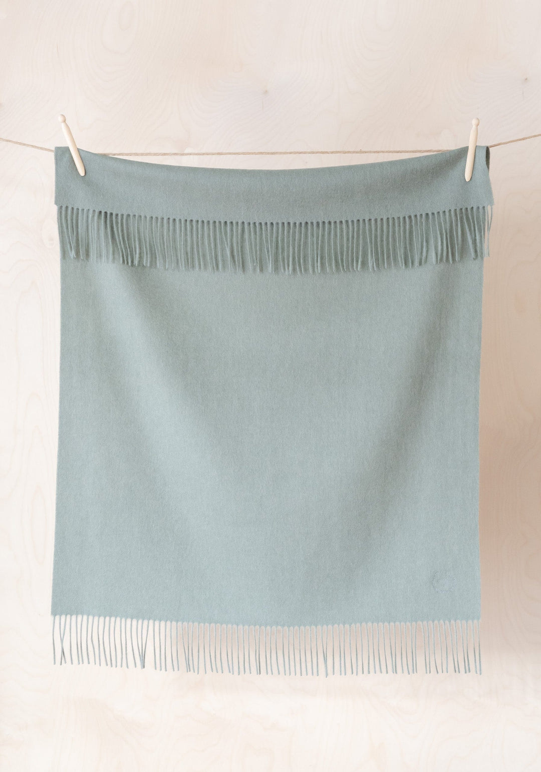 TBCo Lambswool Baby Blanket in Sage Melange hanging on clothesline, featuring soft fringe detail and gentle sage green colour