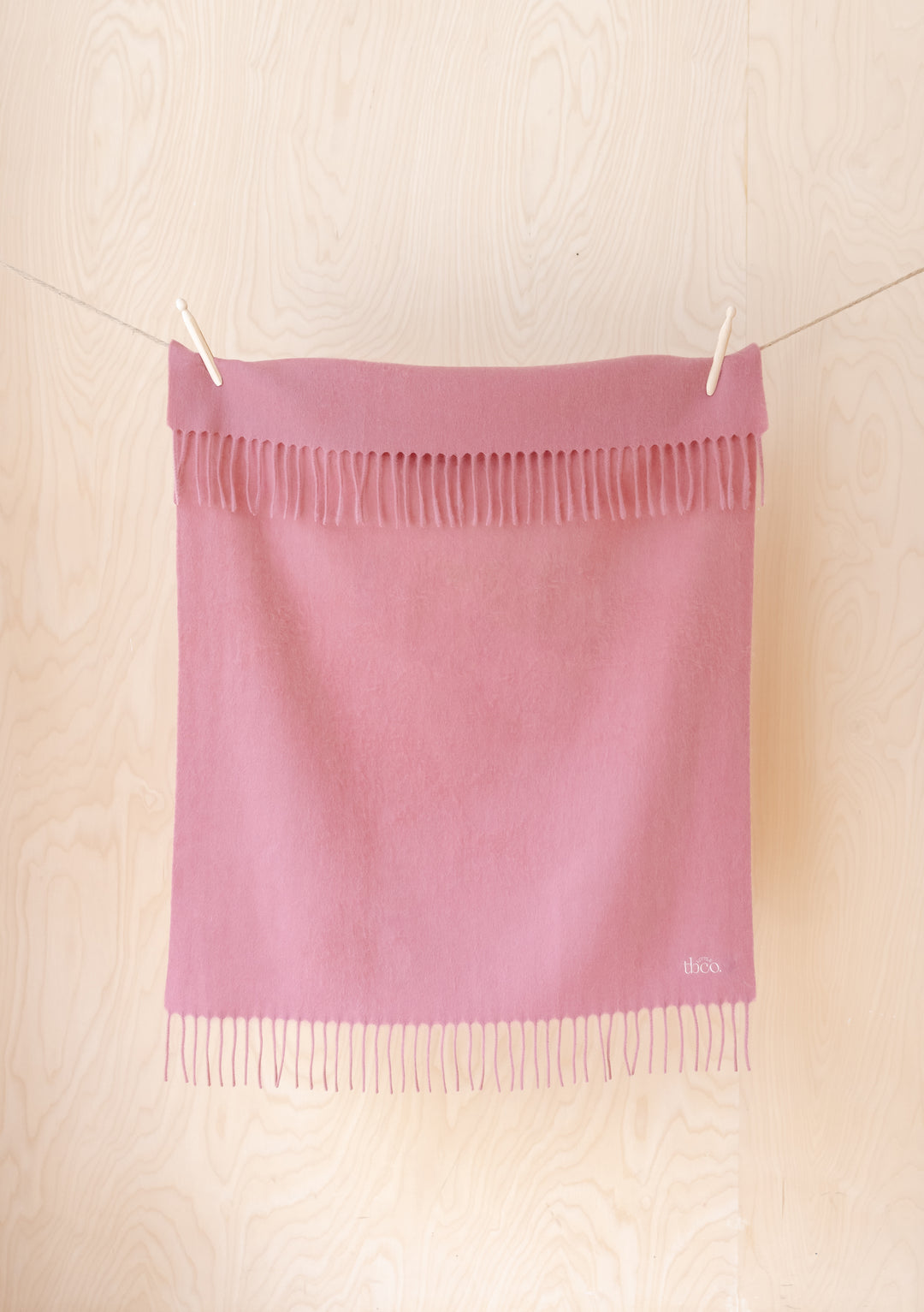 TBCo Lambswool Baby Blanket in Blush hanging from clothesline, displaying fringed edges against light wooden background