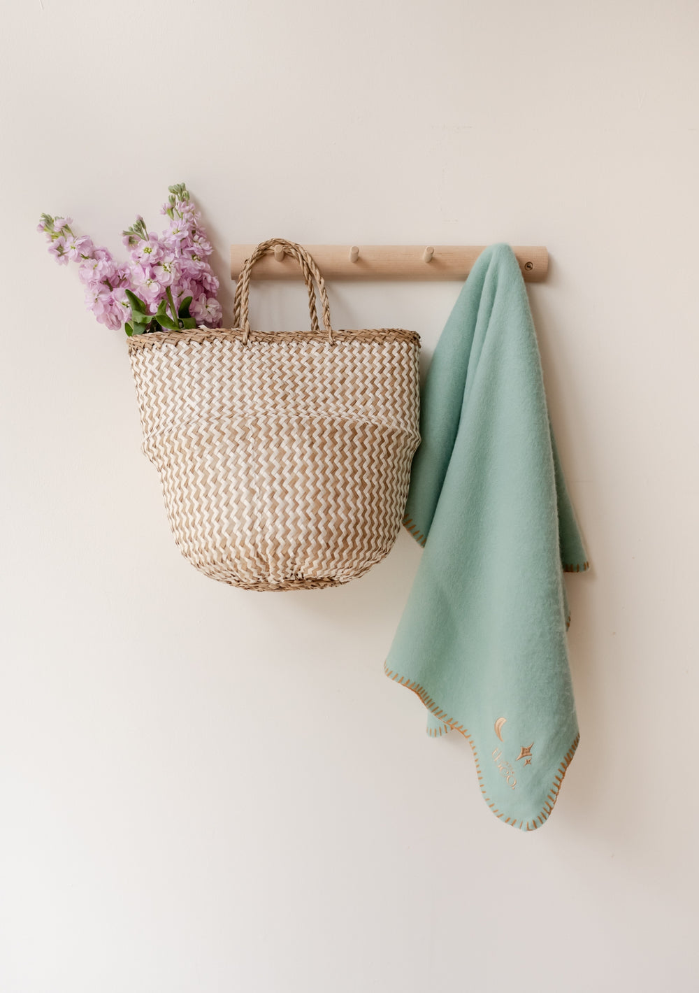 TBCo Lambswool Baby Blanket in mint green with scalloped edges hanging beside woven basket with pink flowers on wooden peg rail