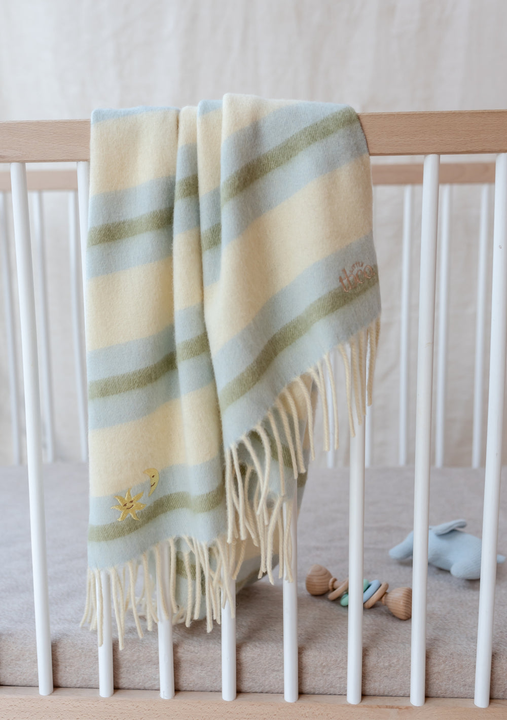 TBCo Lambswool Baby Blanket in blue, green and butter yellow stripes with sun and moon embroidery, draped over white cot rail