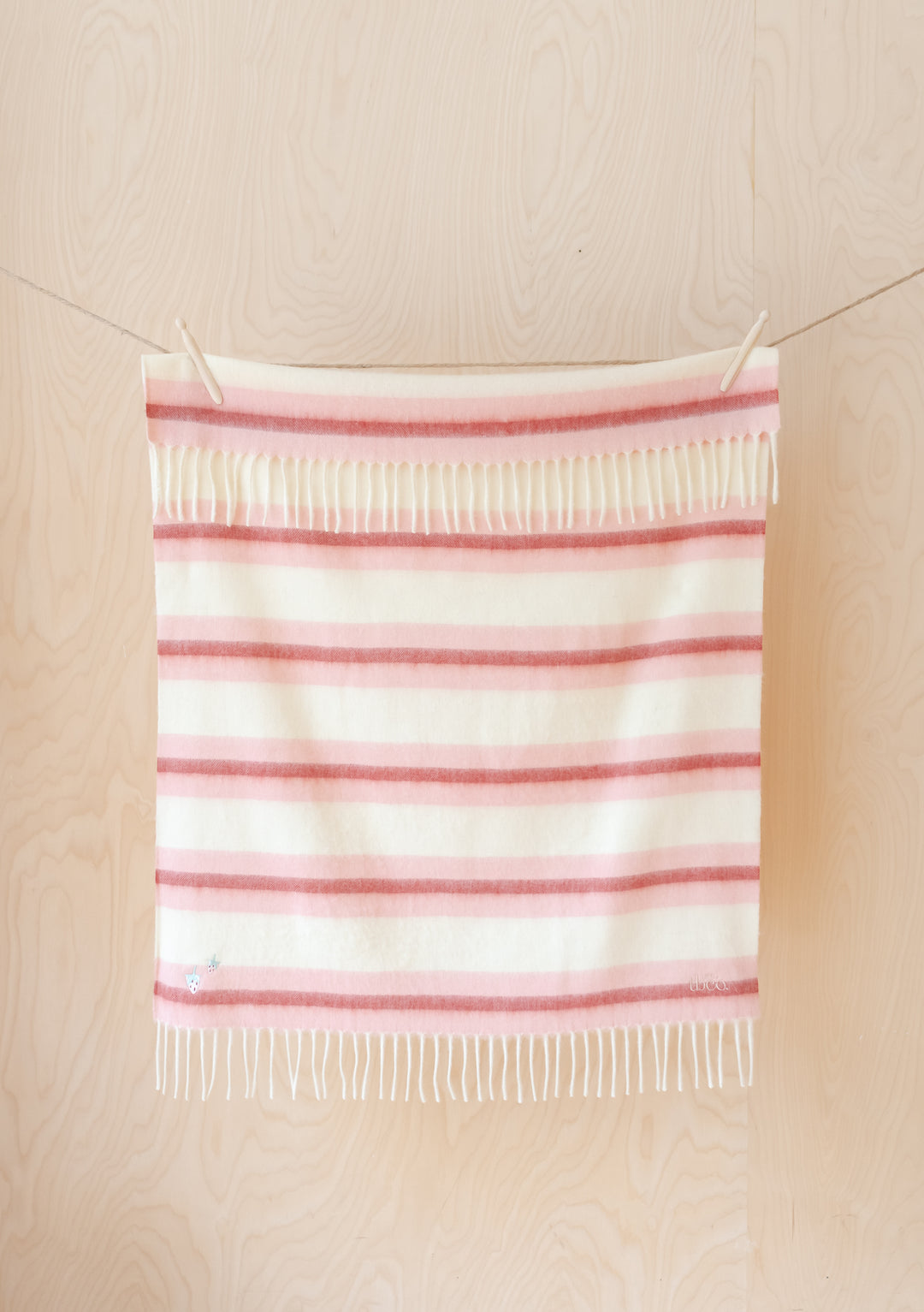 TBCo Lambswool Baby Blanket in Pink Stripe with fringed edges, featuring sun and moon embroidery, displayed on wooden wall