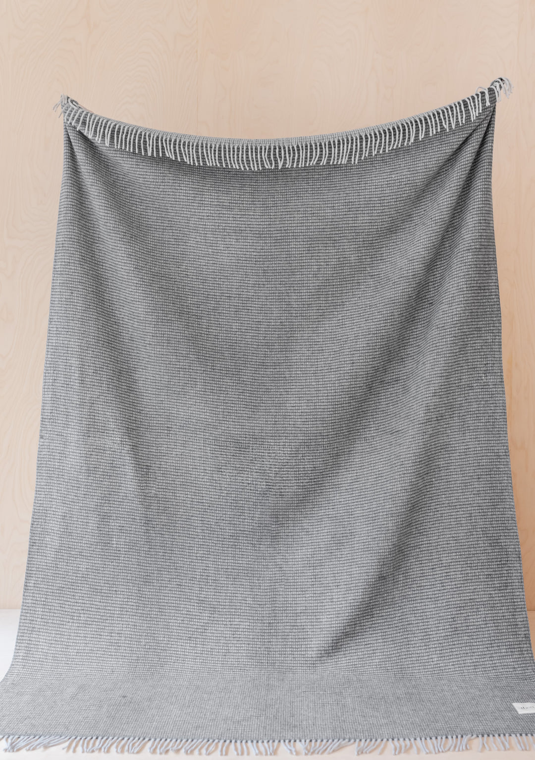 TBCo Recycled Wool Extra Large Blanket in Charcoal Waffle with fringed edges, displayed hanging against beige wall