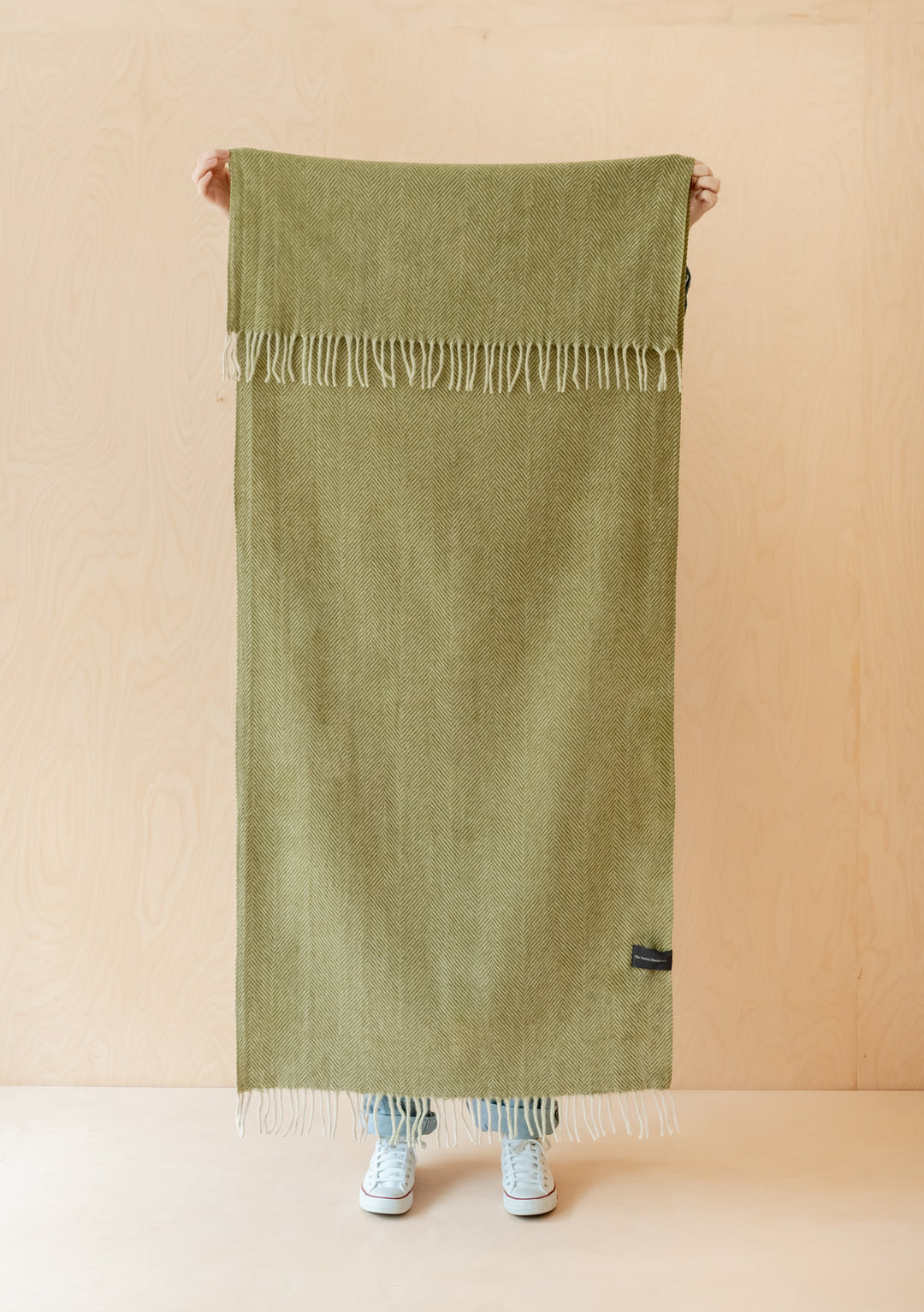 TBCo Recycled Wool Kids Blanket in olive herringbone pattern displayed vertically with fringed edges against beige background