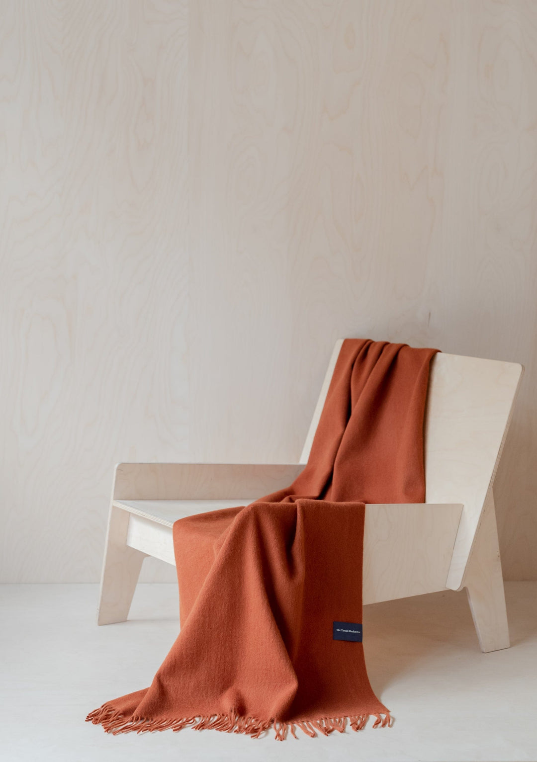 TBCo Lambswool Small Blanket in Rust Melange draped elegantly over a minimalist white chair, showcasing its soft texture and fringe