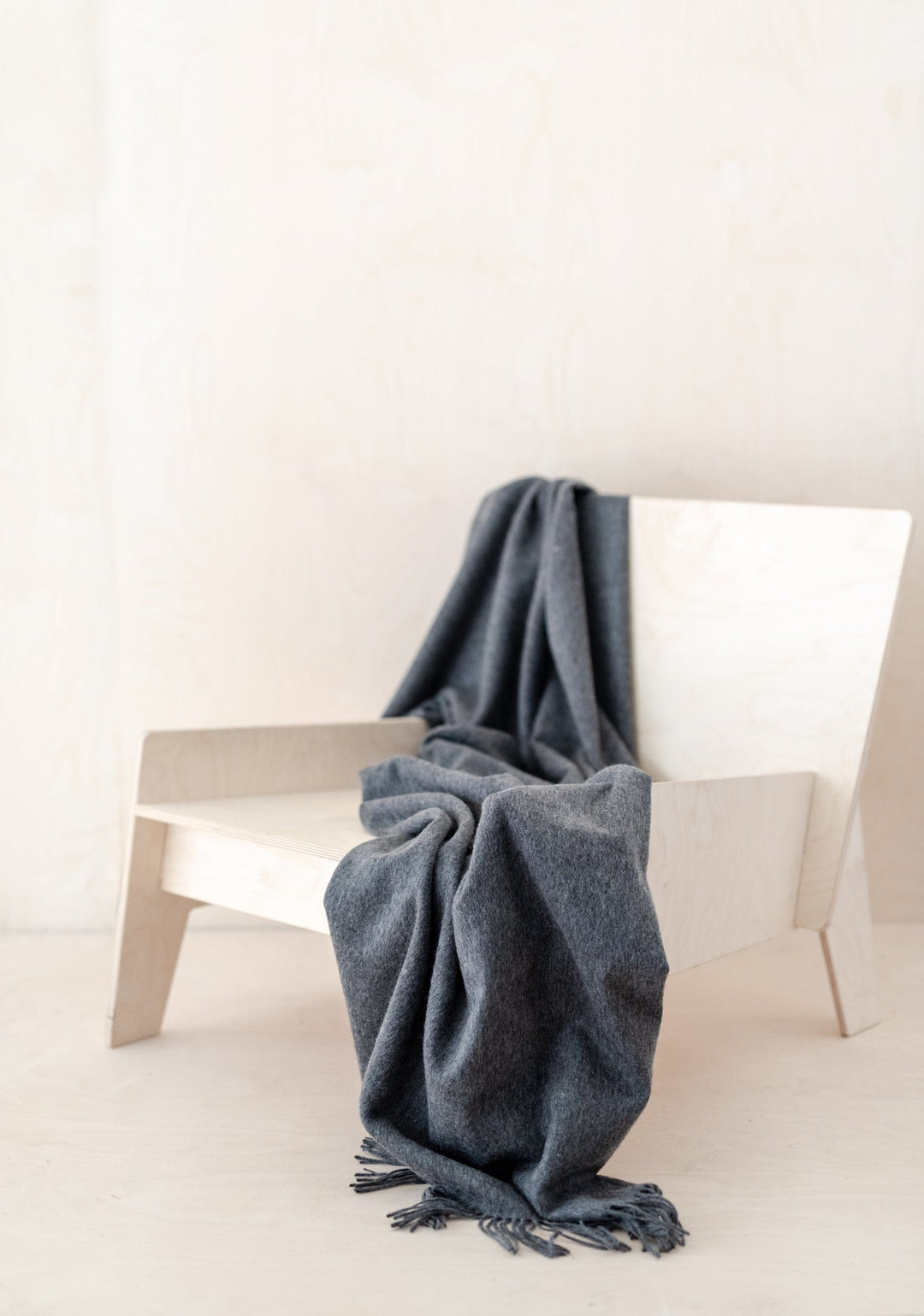 TBCo Lambswool Small Blanket in Charcoal Melange draped elegantly over white modern chair, showcasing soft texture and fringe detail