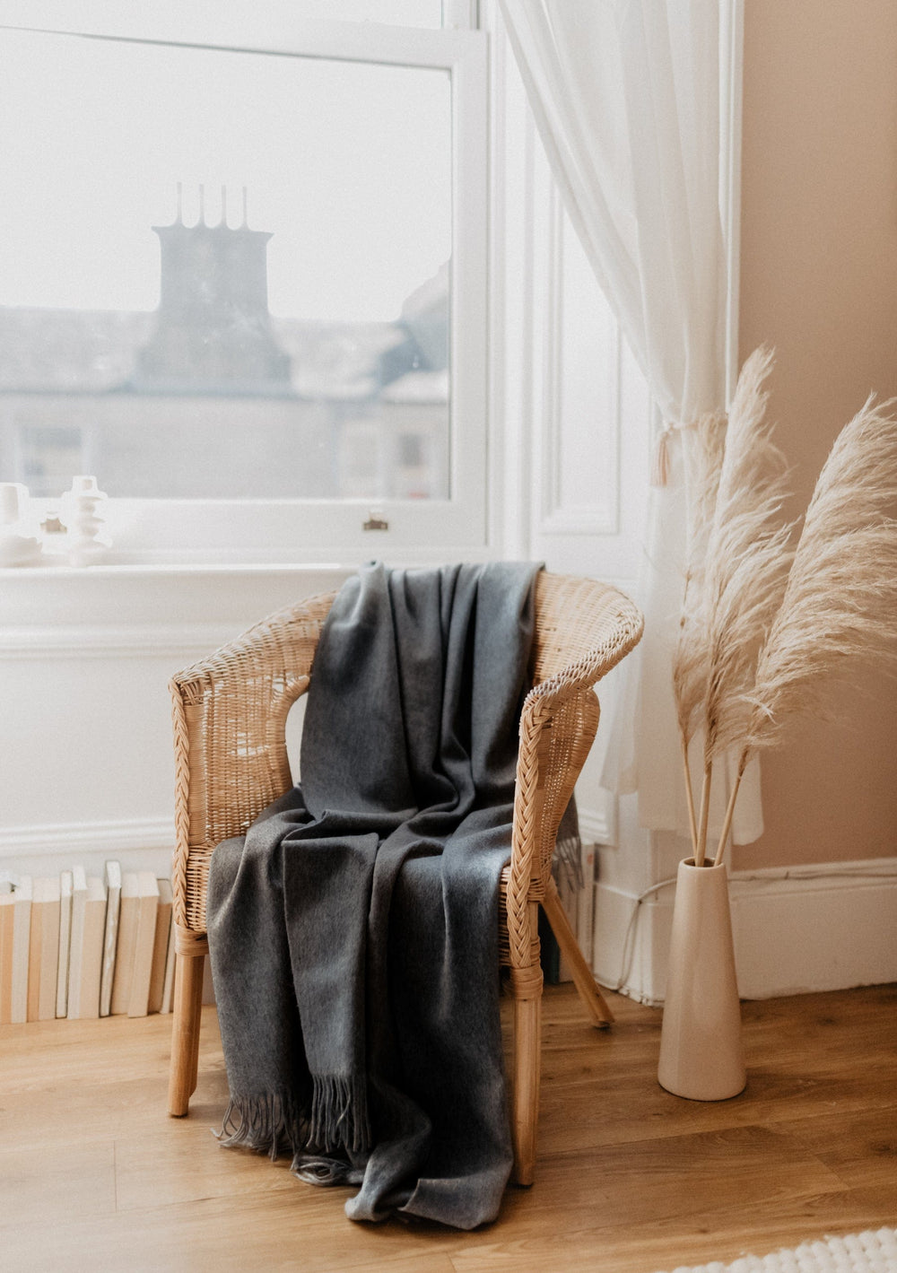 TBCo Lambswool Small Blanket in Charcoal Melange draped elegantly over wicker chair by bright window with pampas grass decor