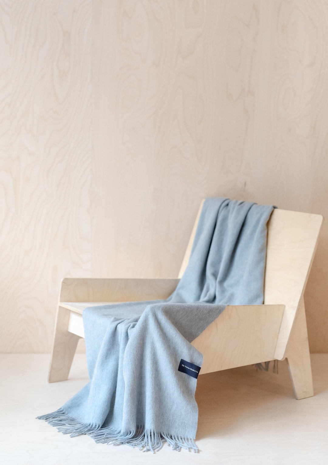 TBCo Lambswool Small Blanket in Light Grey Melange draped elegantly over wooden lounge chair showing texture and fringe detail