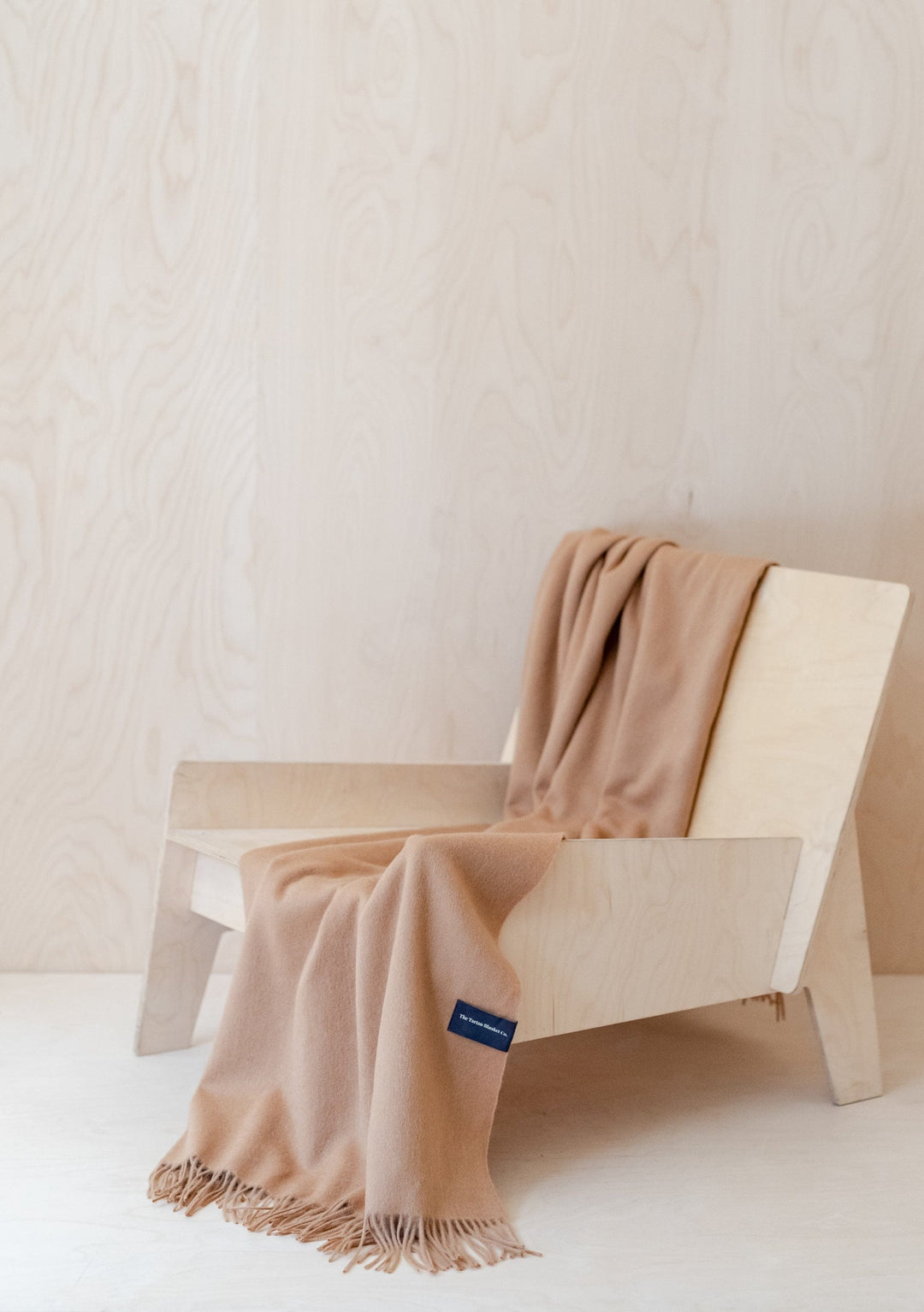 TBCo Lambswool Small Blanket in Camel draped elegantly over a light wooden chair, showcasing its soft texture and fringed edges