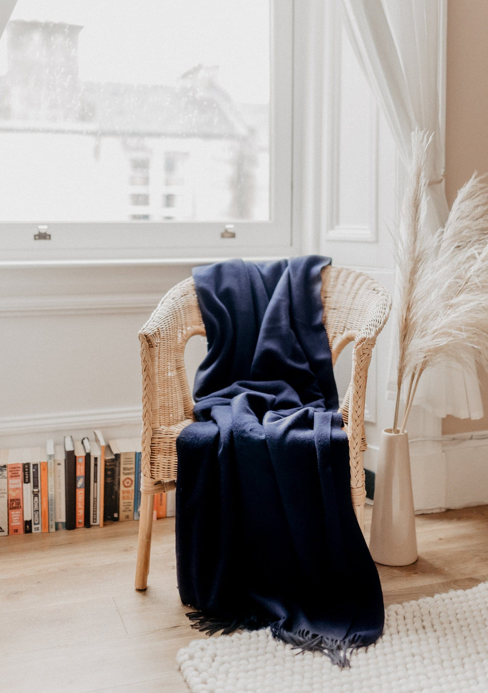 TBCo Lambswool Small Blanket in Navy draped over rattan chair beside window, with pampas grass decor and books