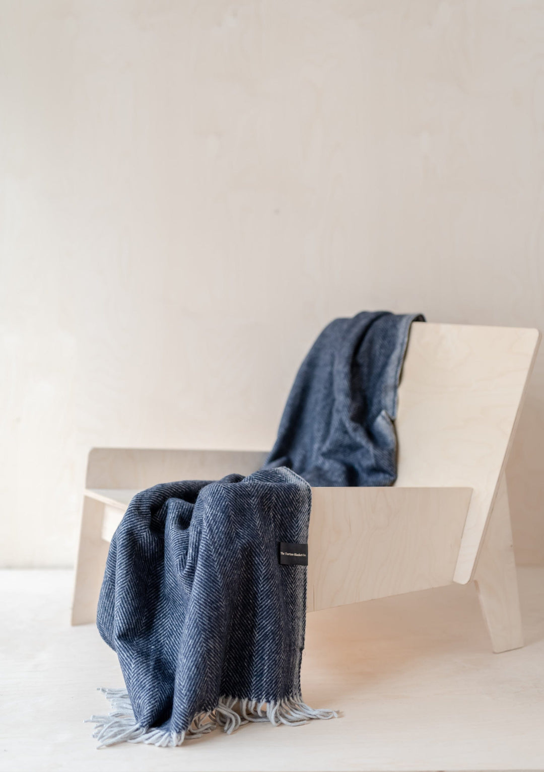 TBCo Recycled Wool Small Blanket in navy herringbone draped elegantly over white chair, showcasing fringed edges and textured weave