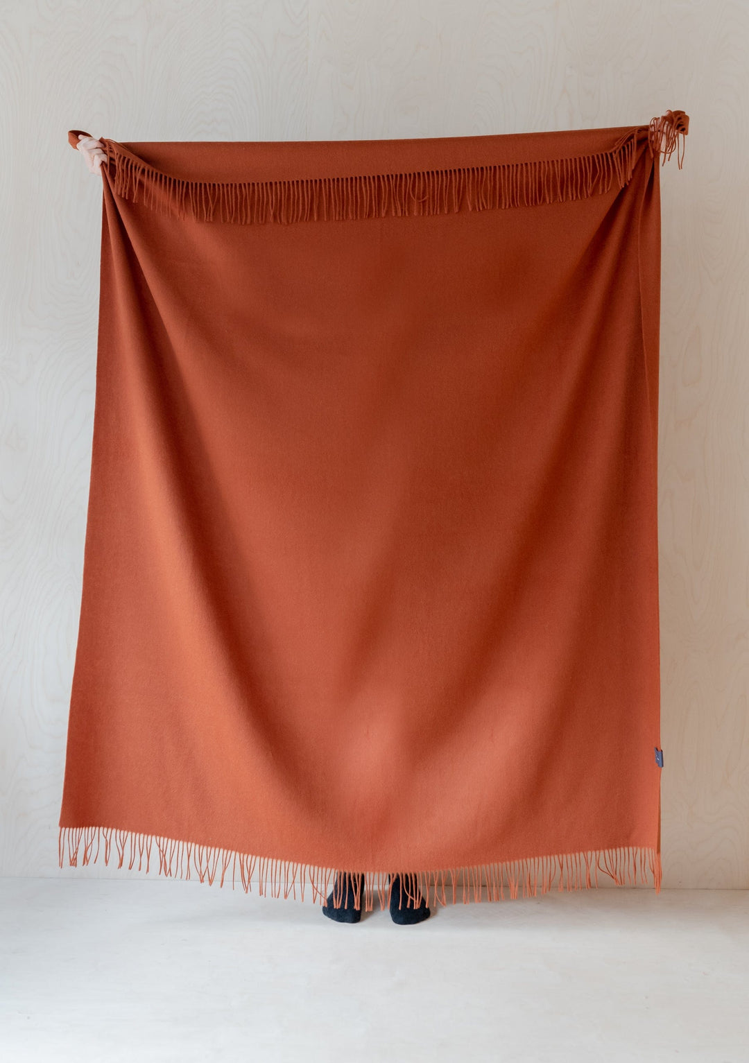 TBCo Lambswool Blanket in Rust Melange with fringed edges displayed against white wall, showcasing luxurious wool texture