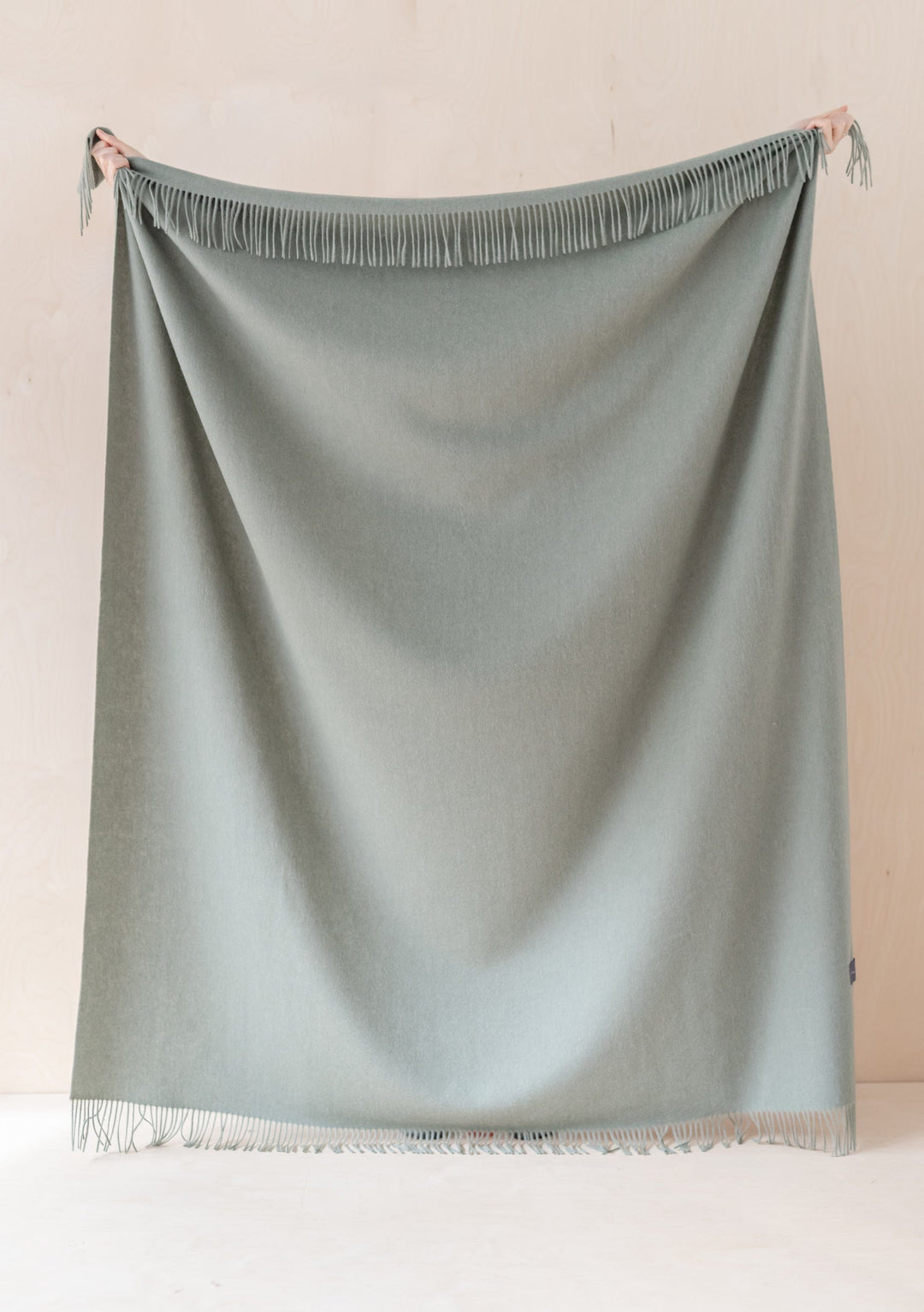 TBCo Lambswool Blanket in Sage Melange with fringed edges, displayed hanging against neutral wall showing soft texture