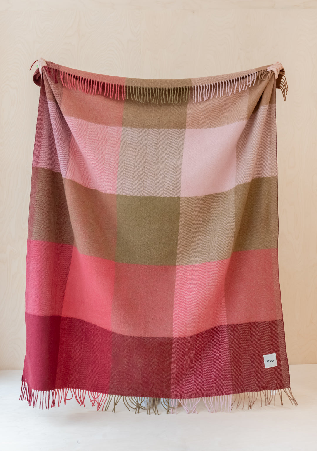 TBCo Recycled Wool Blanket in pink and brown herringbone block check pattern with fringed edges, draped vertically against beige wall