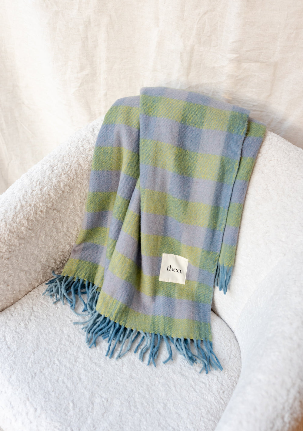 TBCo Recycled Wool Blanket in Green Gingham draped over white chair, showcasing blue and green checkered pattern with fringed edges