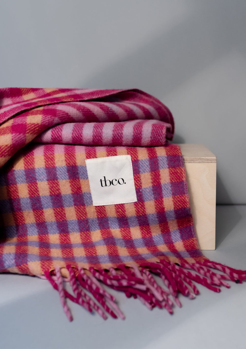 TBCo Recycled Wool Blanket in pink micro gingham pattern with fringed edges, folded on wooden block showing vibrant check design