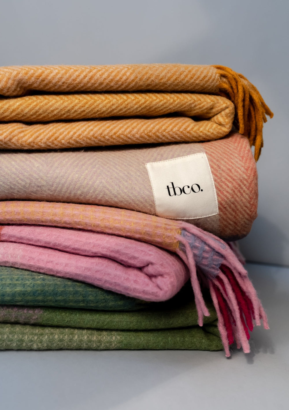 TBCo Recycled Wool Blanket in Green Waffle Block, folded in stack with pink and mustard blankets showing woven texture and brand tag