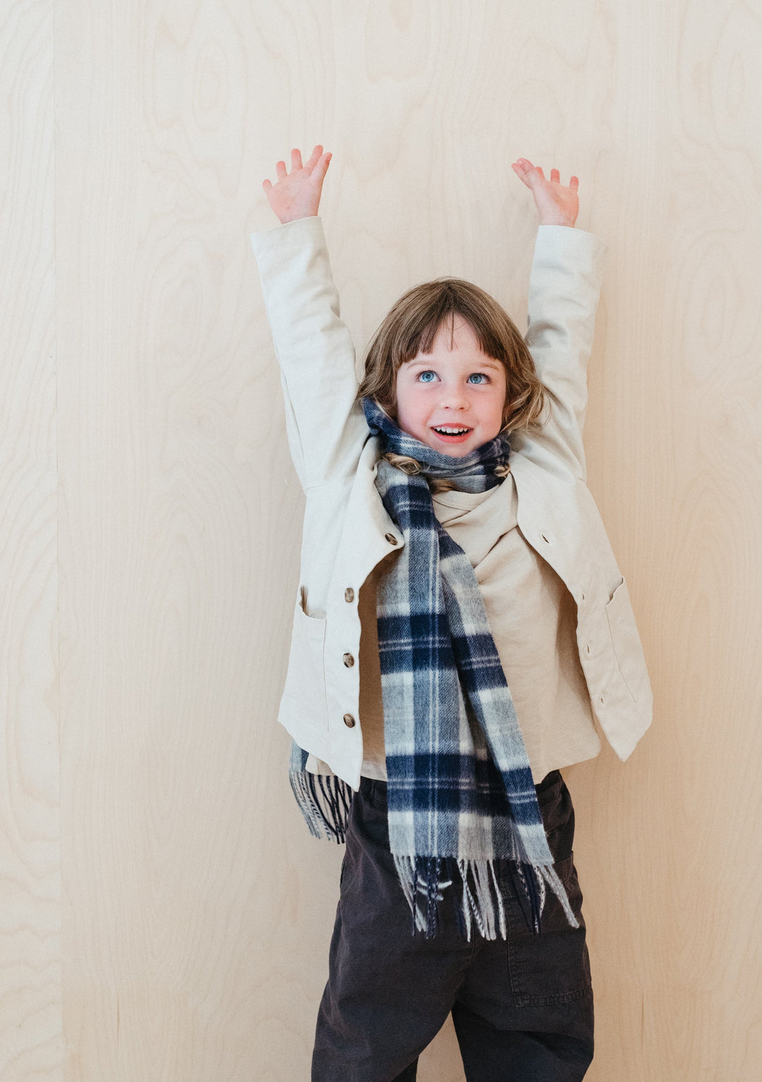 Young child wearing TBCo Lambswool Kids Scarf in Bannockbane Silver Tartan with cream jacket, arms raised playfully against beige wall