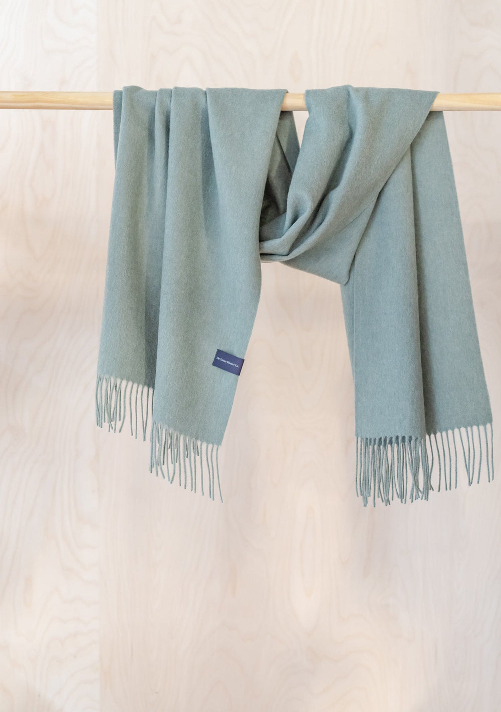 TBCo Lambswool Oversized Scarf in Sage Melange draped elegantly over wooden rail, showcasing fringed edges and soft texture