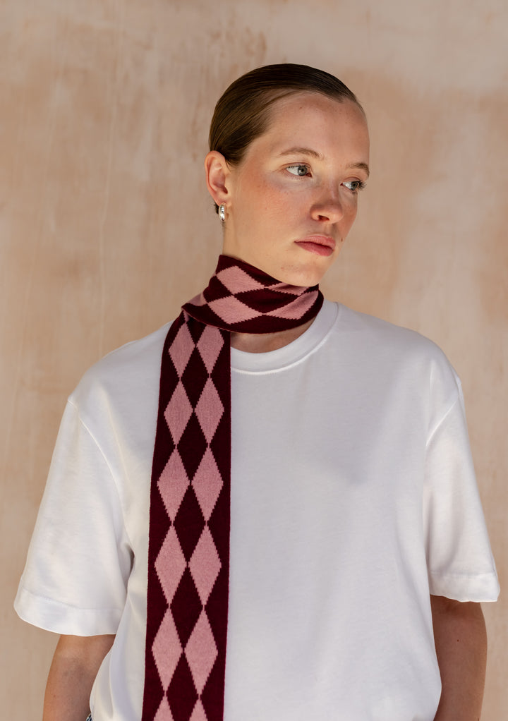 Close-up of a Model wearing a TBCo Merino Wool Skinny Scarf in Burgundy Harlequin elegantly draped around her neck.with a white t-shirt.