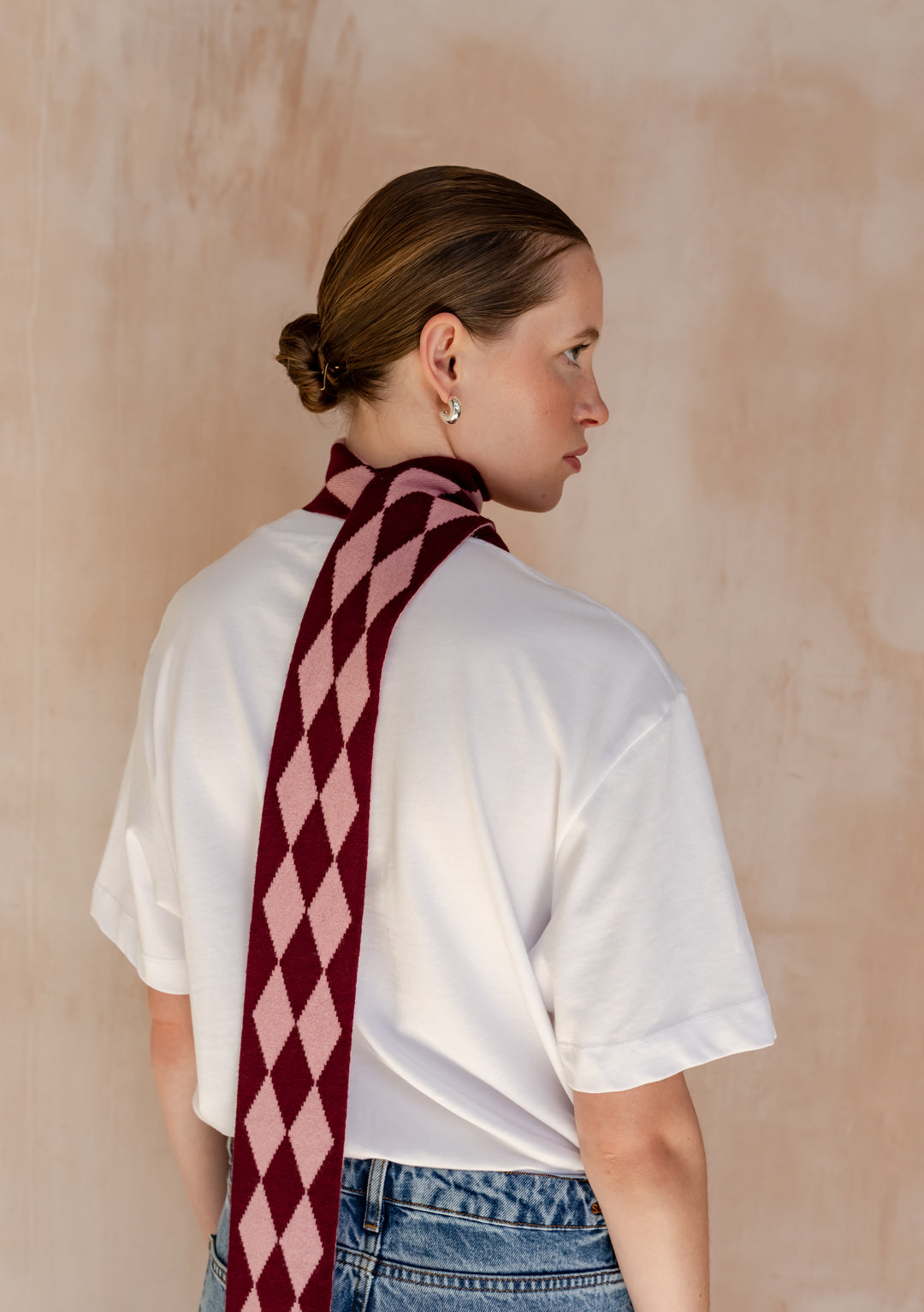 Close-up of a Model from behind wearing a TBCo Merino Wool Skinny Scarf in Burgundy Harlequin elegantly draped around her neck with a white t-shirt.