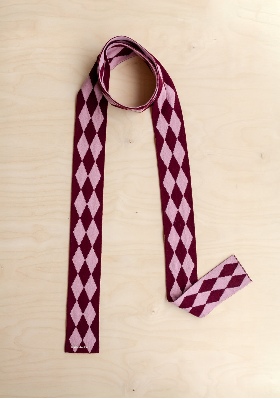 A TBCo Merino Wool Skinny Scarf in Burgundy Harlequin lying on a wooden background. 