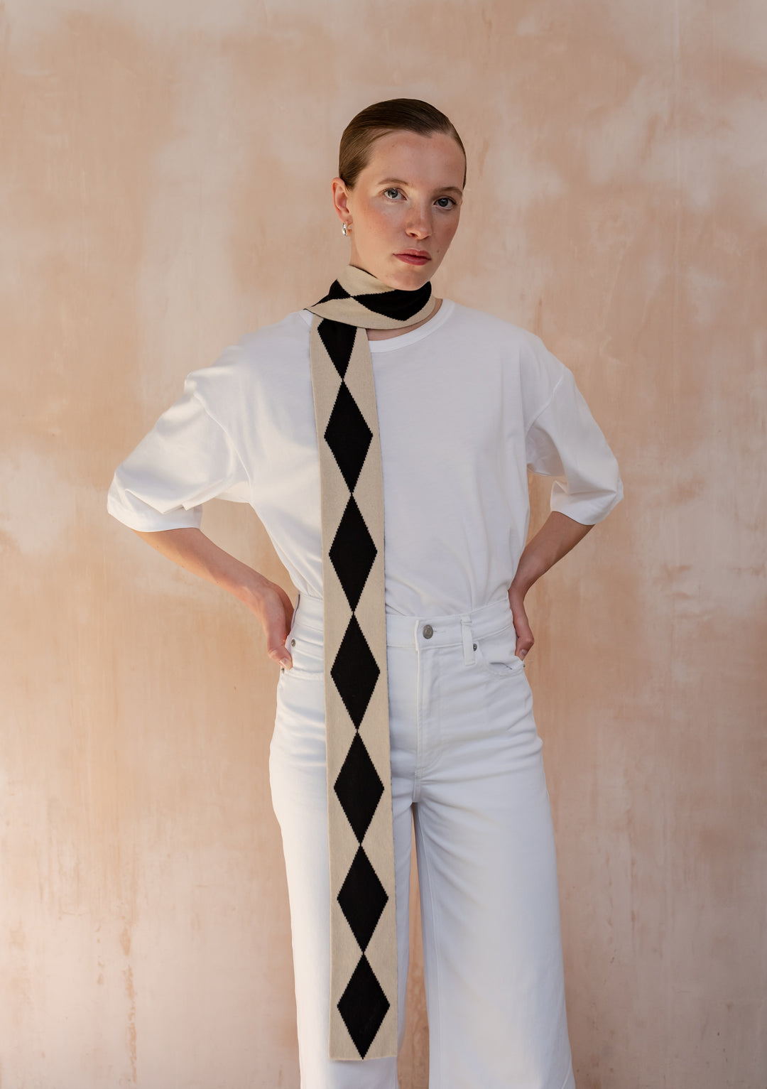A Model wearing a TBCo Merino Wool Skinny Scarf in Black Harlequin elegantly draped around her neck with a white t-shirt and white jeans.