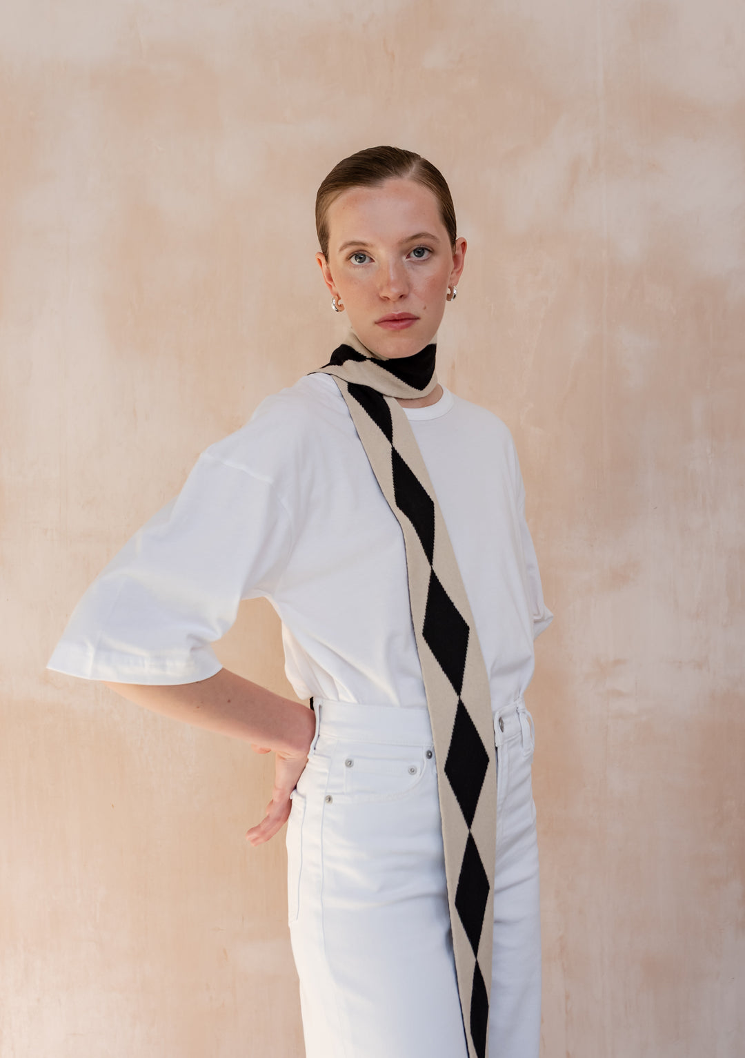 A Model wearing a TBCo Merino Wool Skinny Scarf in Black Harlequin elegantly draped around her neck with a white t-shirt and white jeans.