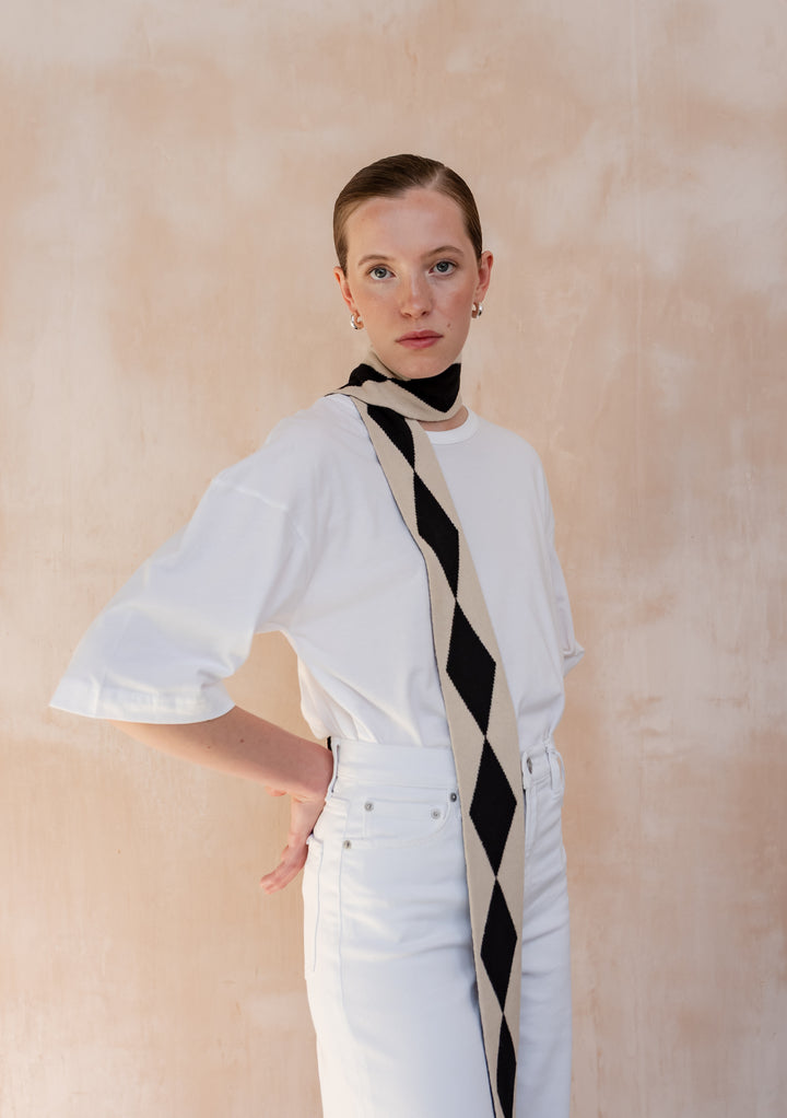 A Model wearing a TBCo Merino Wool Skinny Scarf in Black Harlequin elegantly draped around her neck with a white t-shirt and white jeans.